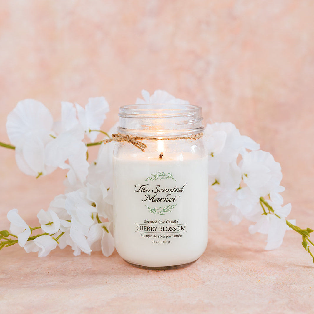 Candle in a mason jar with 'The Scented Market' label, surrounded by cherry blossom branches on a soft pink background.