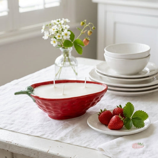 Red strawberry shaped bowl filled with a 3 wick dye free 100% soy wax candle and strawberries on a white tablecloth in a bright room.