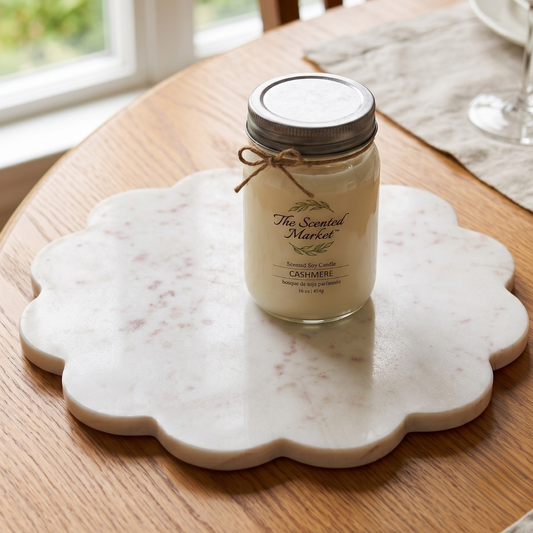 A jar candle from The Scented Market in a clear glass mason jar with a metal lid and twine bow, placed on a scalloped white marble tray on a wooden table near a window.