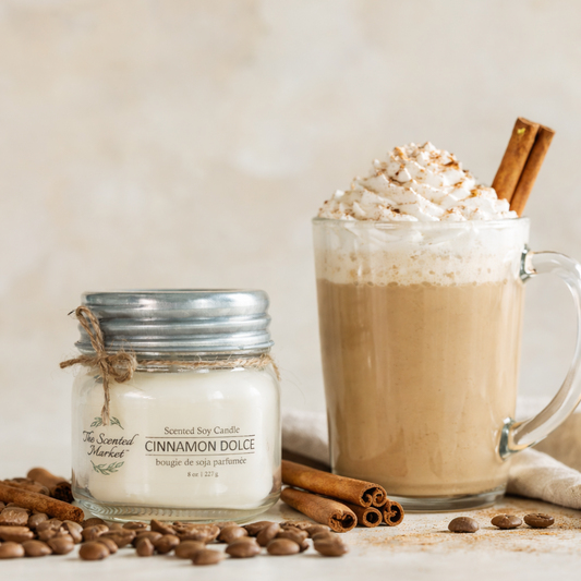 Cinnamon Dolce candle next to a mug of hot chocolate with whipped cream and a cinnamon stick on a wooden surface.