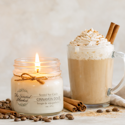 Candle labeled 'The Scented Market Cinnamon Dolce' next to a mug of hot chocolate with whipped cream and a cinnamon stick.