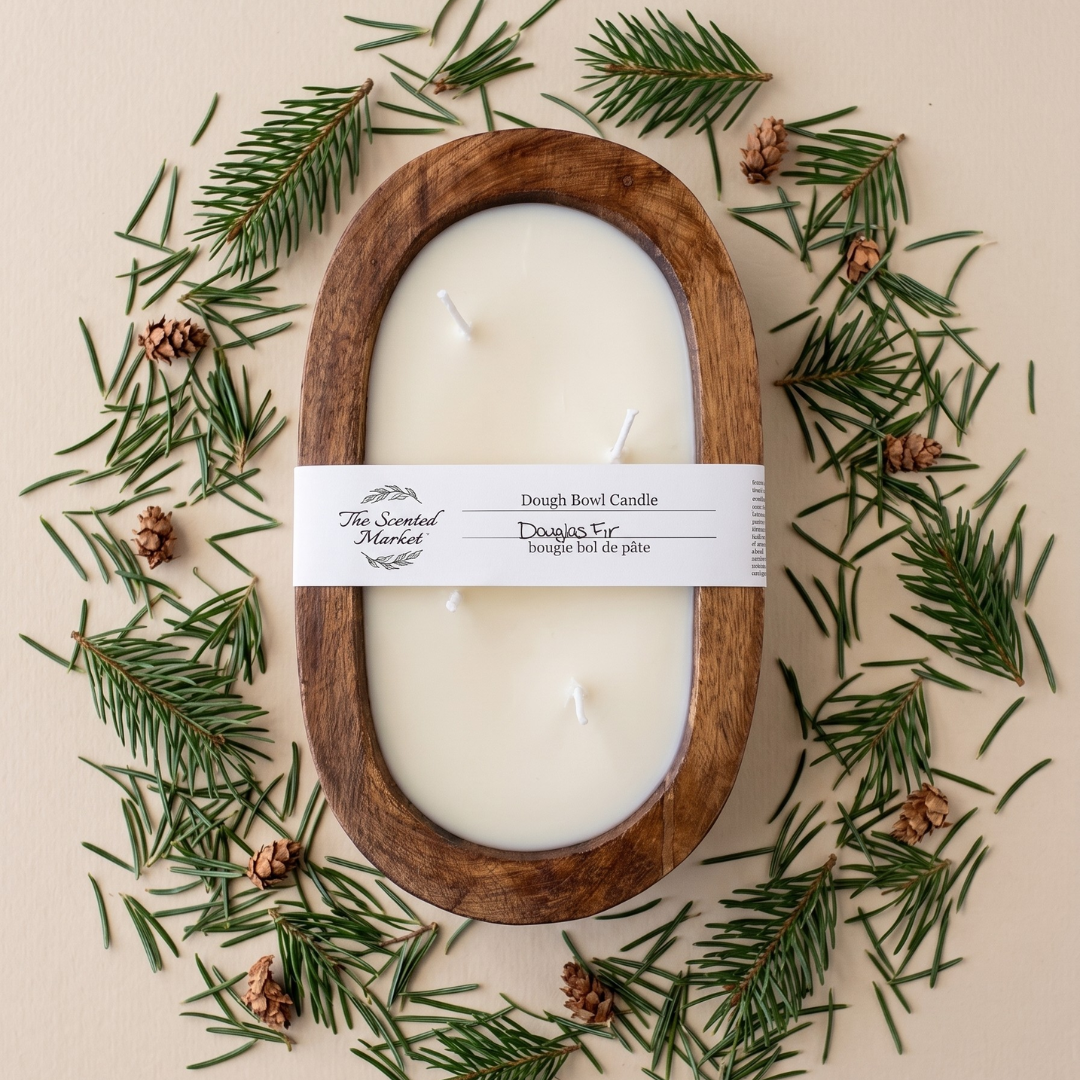 A wooden oval candle holder filled with white wax, wrapped with a white paper label reading 'The Sacred Melt' and 'Dough Bread Candle'. The candle is arranged in a flat lay on a beige background, surrounded by pine needles and small pinecones.