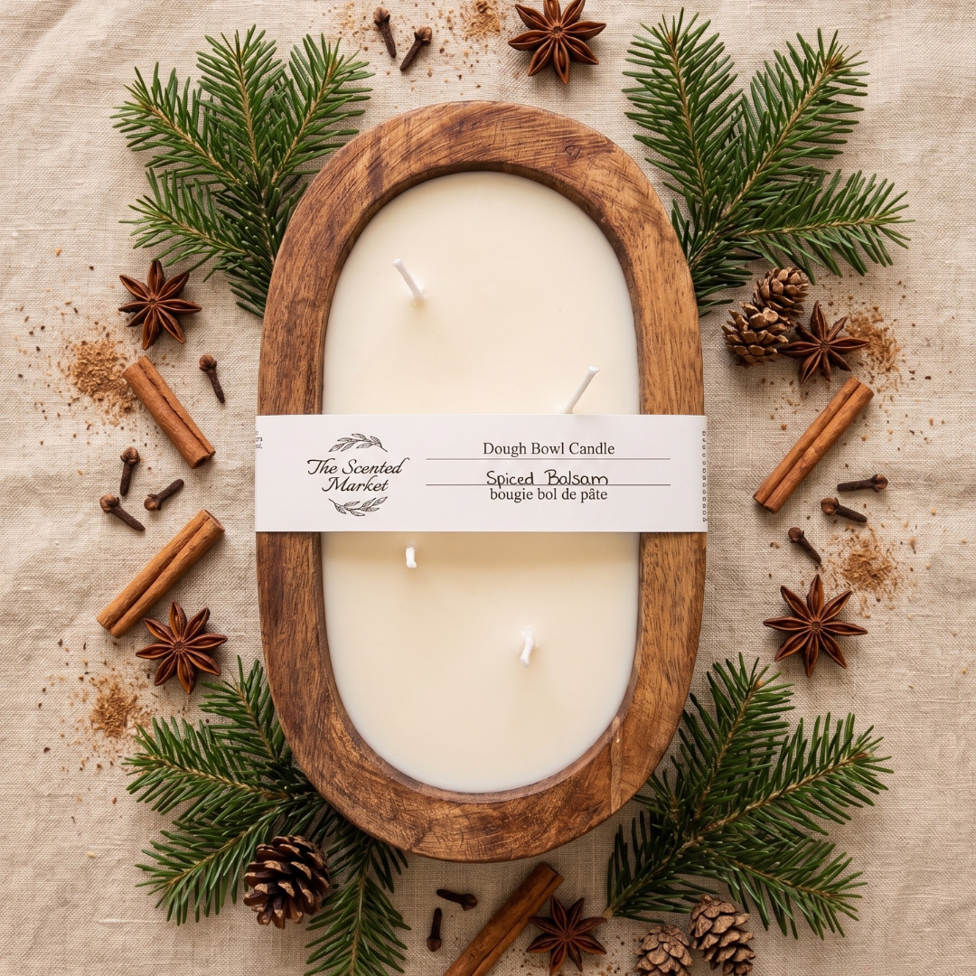 An oval wooden candle holder filled with cream-colored wax, wrapped with a white paper band labeled 'The Scented Market Dough Bowl Candle.' The candle is arranged in a styled flat lay on a beige fabric background, surrounded by green pine branches, star anise, cinnamon sticks, cloves, and pinecones.